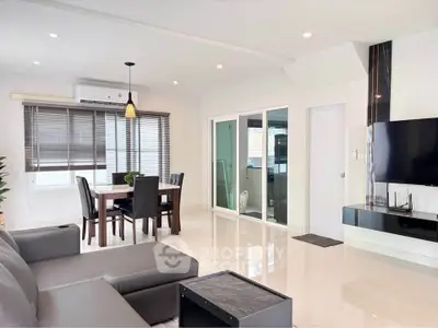 Modern living room with sleek furniture and dining area, featuring a wall-mounted TV and large windows.
