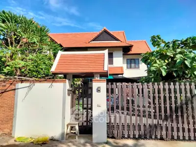 Charming two-story house with red-tiled roof and gated entrance, surrounded by lush greenery.