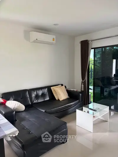 Modern living room with black leather sofa, glass coffee table, and large window overlooking garden.