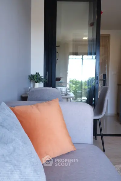 Modern living room with stylish gray sofa and orange cushion, featuring glass doors leading to a bright dining area.
