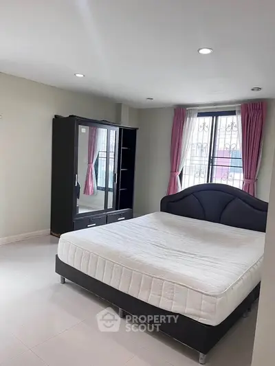 Spacious bedroom with modern bed and wardrobe, featuring large window with pink curtains.