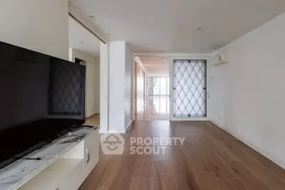 Spacious modern living room with wooden flooring and large TV, leading to a bright hallway.