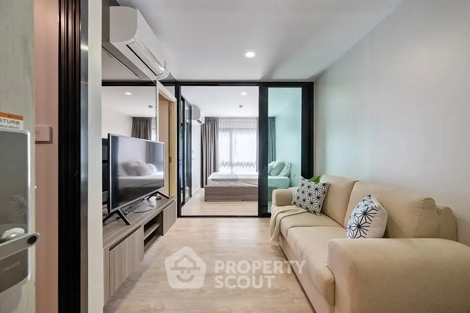 Modern living room with beige sofa and TV, leading to a cozy bedroom through glass doors.