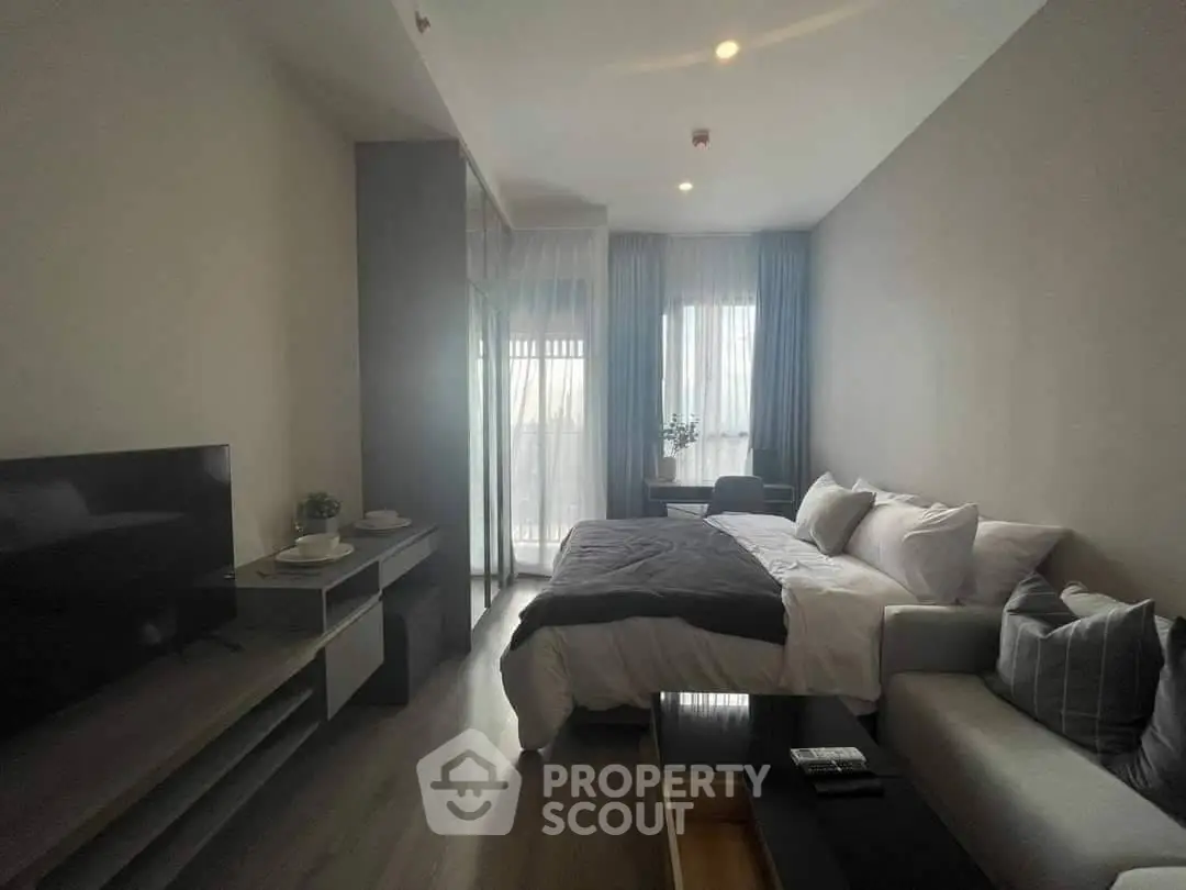 Modern studio apartment with cozy bed, sleek TV setup, and natural light from large windows.