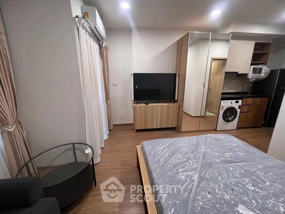 Modern studio apartment with wooden flooring, featuring a bed, TV, and washing machine in an open layout.