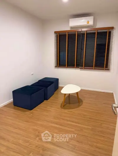 Minimalist room with wooden flooring, blue ottomans, and a small white table under an air conditioner.