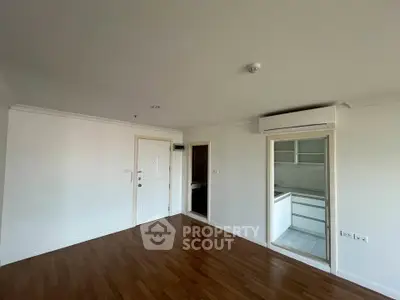 Spacious empty room with wooden flooring and air conditioning, adjacent to a modern kitchen.