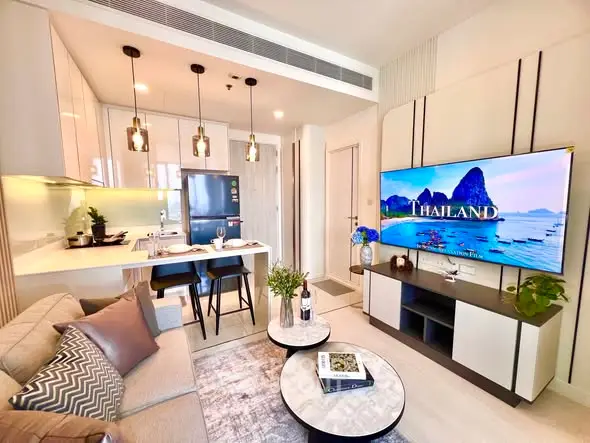 Modern living room with open kitchen, stylish decor, and large TV displaying Thailand scenery.