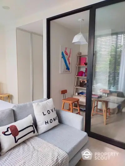 Modern living space with chic decor, featuring a cozy grey sofa adorned with stylish cushions, sleek glass partition leading to a bright study room with vibrant shelves and art.