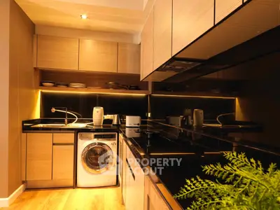 Modern kitchen with sleek black countertops and built-in appliances, featuring a washing machine and stylish lighting.