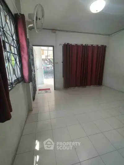 Spacious living room with tiled floor and large windows, featuring red curtains and a ceiling fan.