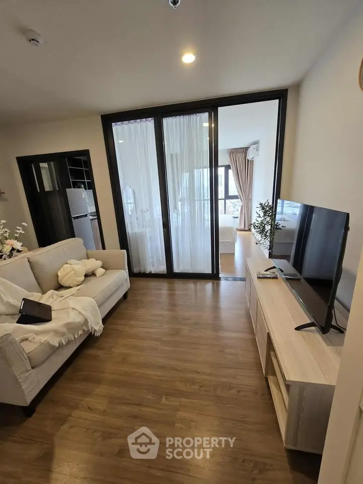 Modern living room with wooden flooring, cozy sofa, and TV unit, leading to a bright bedroom with large windows.