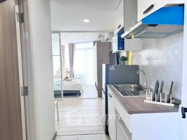 Modern apartment kitchen with open layout leading to a bright bedroom with large windows.