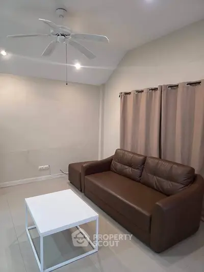 Modern living room with brown leather sofa and white coffee table, featuring ceiling fan and neutral curtains.