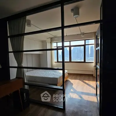 Modern bedroom with large windows and city view, featuring sleek glass partition and wooden flooring.
