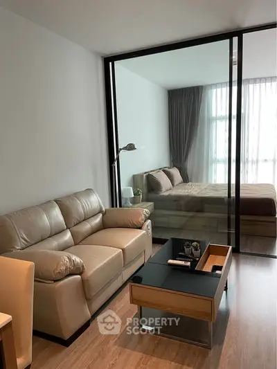 Modern living room with glass partition leading to a cozy bedroom, featuring stylish furniture and natural light.
