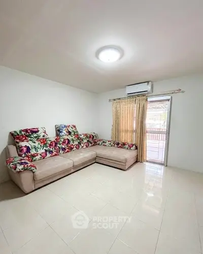 Spacious living room with floral sofa and air conditioning, featuring natural light from large window.