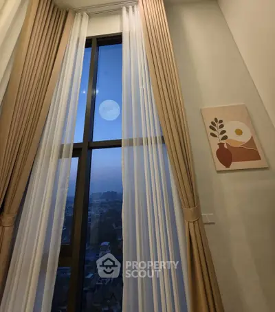 Elegant high-rise window view with stylish curtains and modern wall art, showcasing cityscape and moonlit sky.