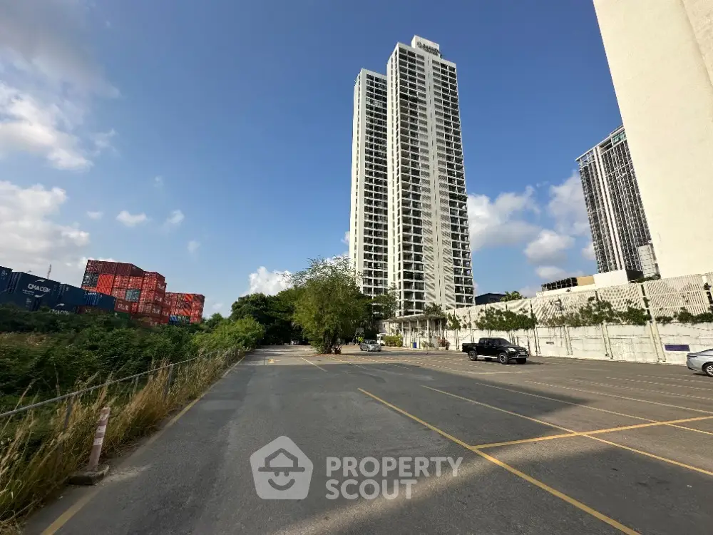 Majestic high-rise condominium towers soar into the blue sky, flanked by lush greenery and a spacious parking area, offering a blend of urban living and nature.