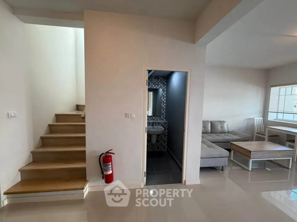 Modern living room with staircase and adjacent bathroom, featuring sleek furniture and natural light.