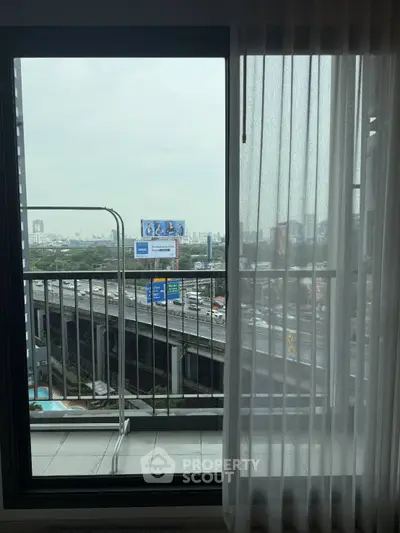 City view from a modern apartment balcony with glass door and sheer curtains.