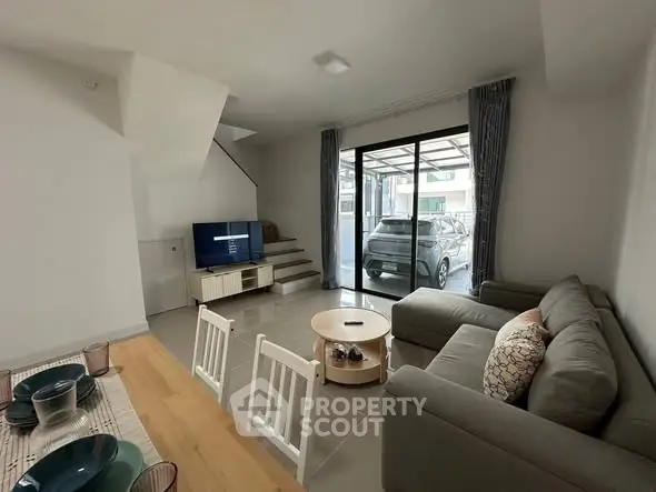 Modern living room with cozy sofa, TV, and dining area, featuring a view of the carport through glass doors.