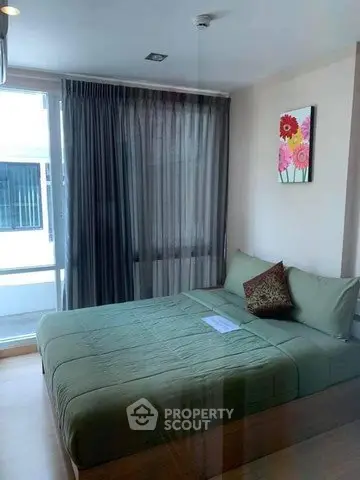 Cozy bedroom with green bedding and floral artwork, featuring large window with sheer curtains.