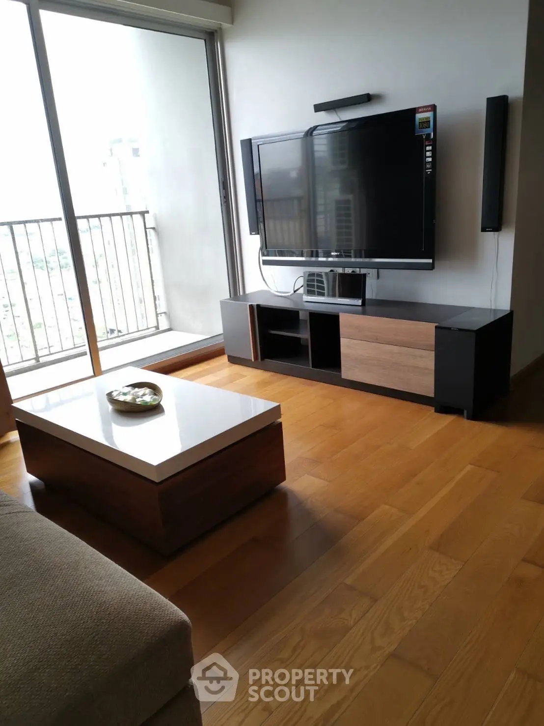 Modern living room with wooden flooring and large TV, featuring a sleek coffee table and balcony view.