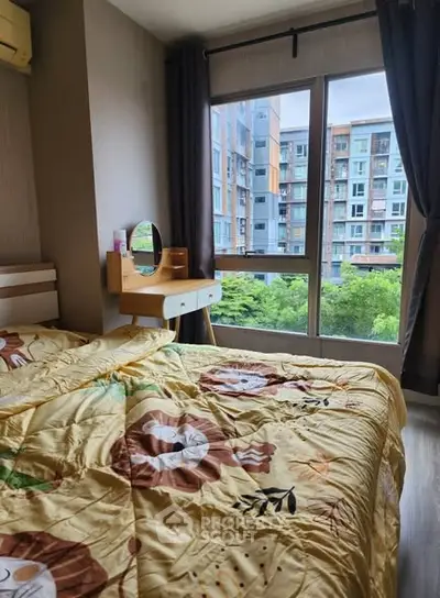 Cozy bedroom with large window and vibrant bedding overlooking lush greenery and modern buildings.