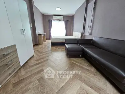 Spacious modern studio apartment with sleek herringbone wood flooring, plush leather sofa, and a cozy sleeping area bathed in natural light from the large window, perfect for urban living.
