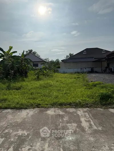 Spacious residential plot with lush greenery and modern houses under a clear sky.