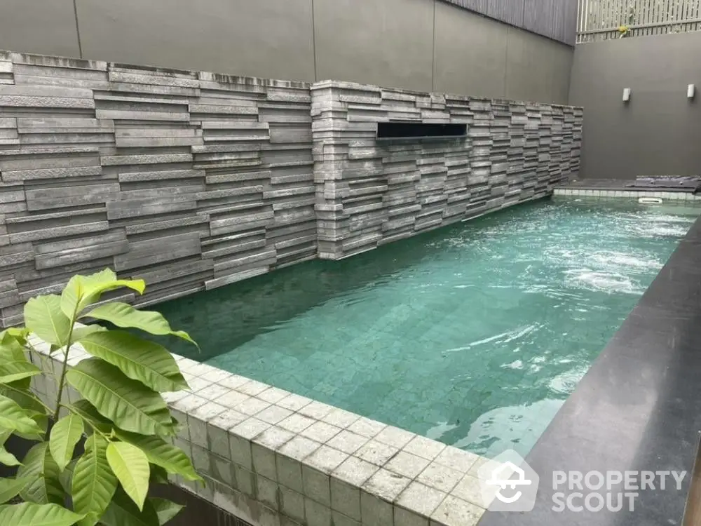 Inviting modern pool with stylish stone wall design, surrounded by serene grey tones and lush greenery, perfect for urban relaxation.