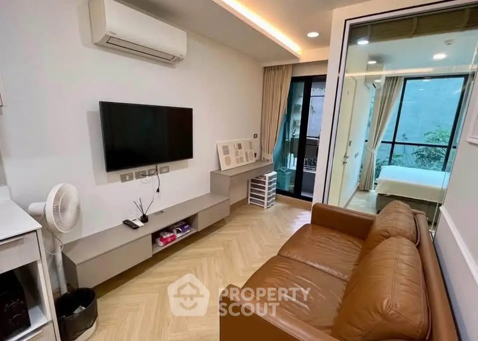Modern living room with brown leather sofa, wall-mounted TV, and air conditioning, leading to a bedroom with balcony view.