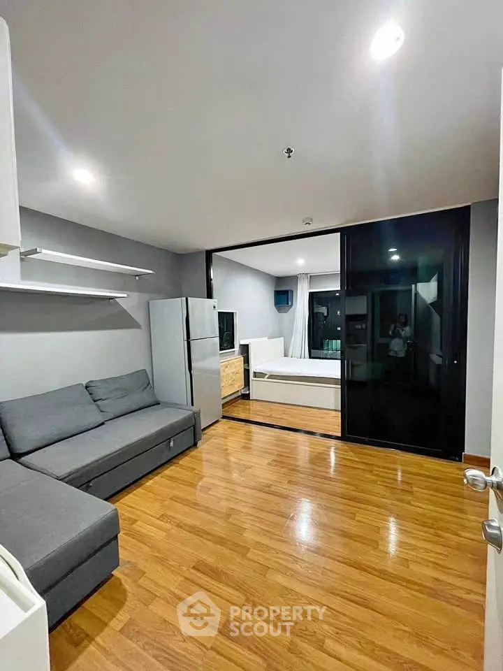 Modern living room with sleek wooden flooring and open layout, featuring a cozy gray sofa and a spacious fridge.
