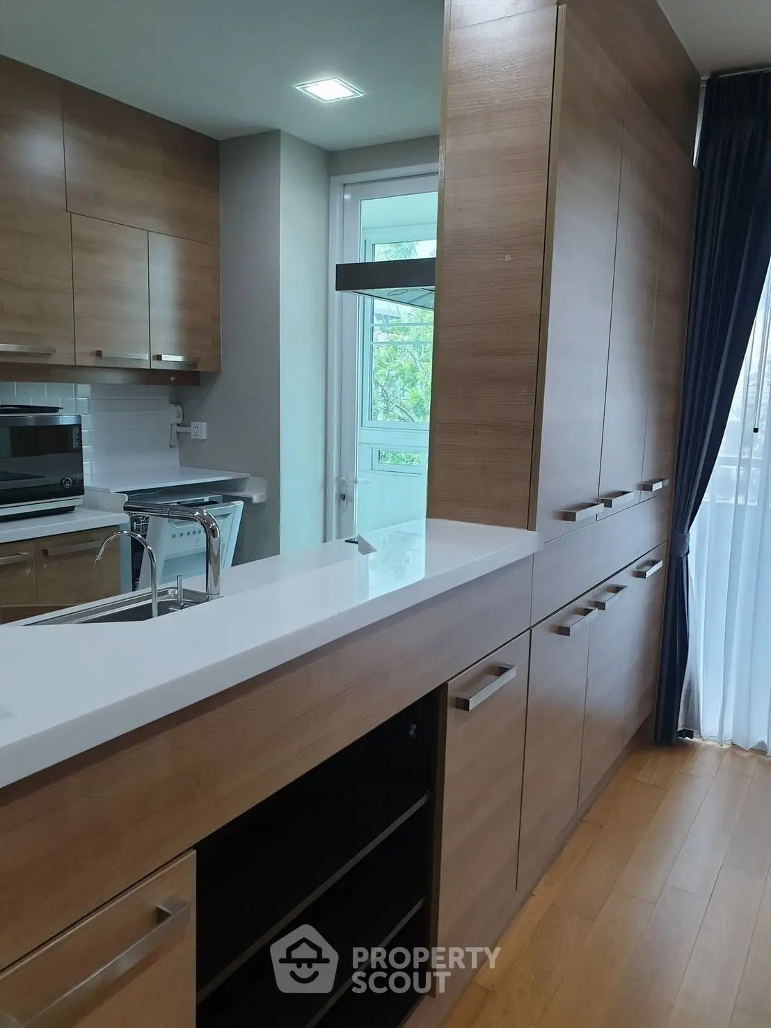 Modern kitchen with sleek wooden cabinets and white countertops, featuring a built-in oven and large window.