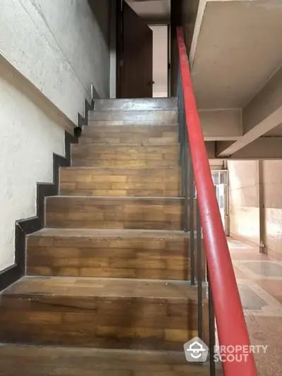 Elegant wooden staircase with a striking red handrail, leading to upper levels in a modern residential building, showcasing architectural detail and design.