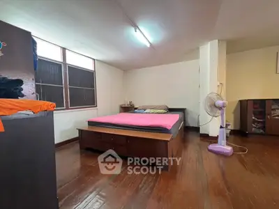 Spacious bedroom with wooden flooring and natural light, featuring a cozy bed and fan.