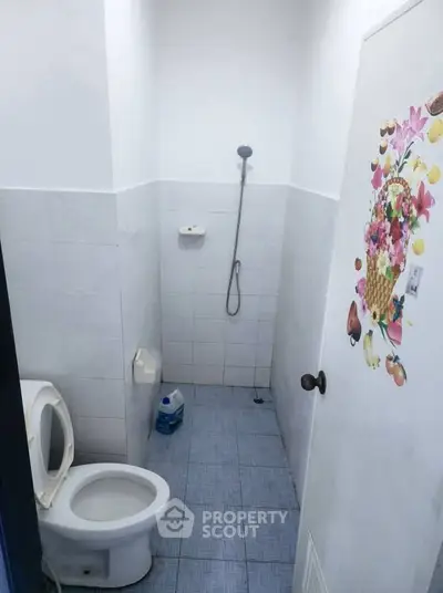 Compact bathroom with shower and toilet, featuring simple white tiles and a decorative door sticker.