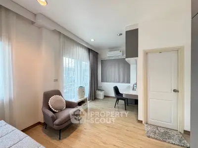 Modern bedroom with cozy seating and workspace, featuring large windows and wooden flooring.