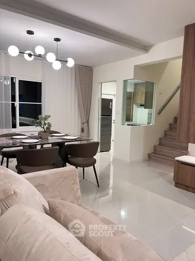 Modern open-plan living room with elegant dining area and sleek staircase, featuring contemporary lighting and neutral decor.