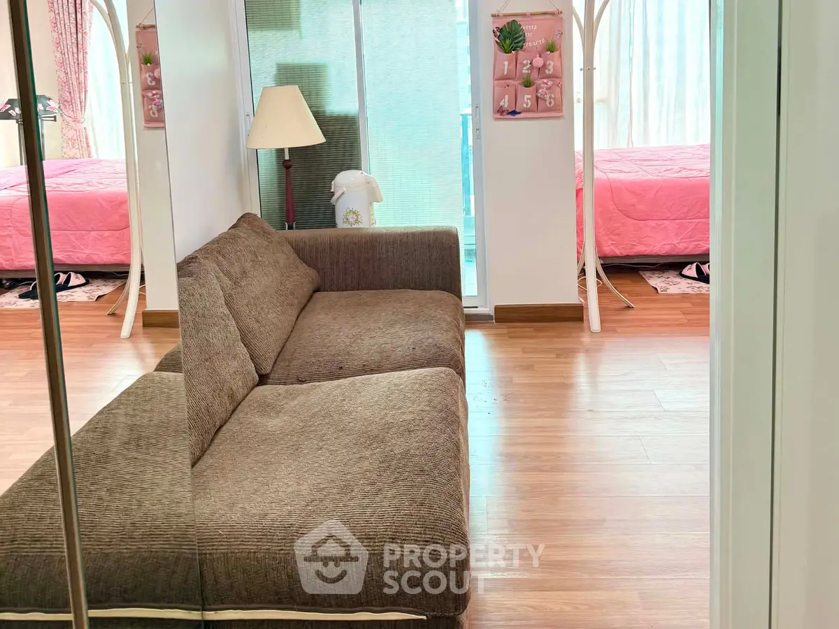 Cozy living room with brown sofa and mirrored closet, leading to a bright bedroom with pink bedding.