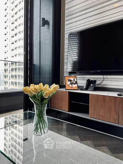 Modern living room with sleek TV setup and glass coffee table, featuring a vase of yellow tulips and city view.