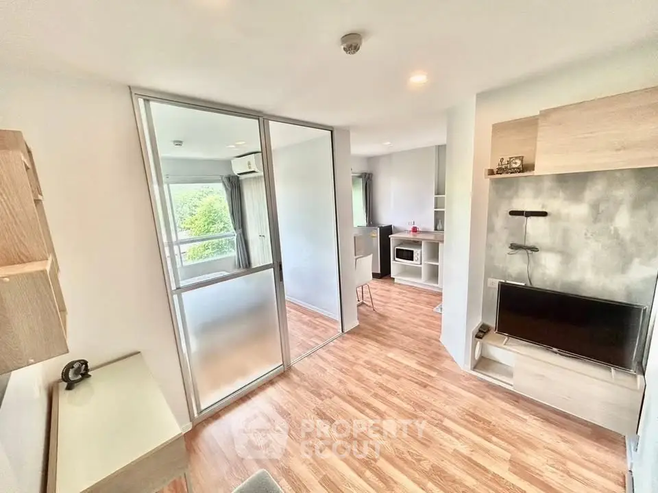 Modern living room with wooden flooring and glass partition, featuring a cozy TV setup and natural light.