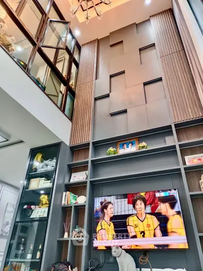 Spacious living room with high ceiling and modern decor, featuring a large TV and elegant shelving.