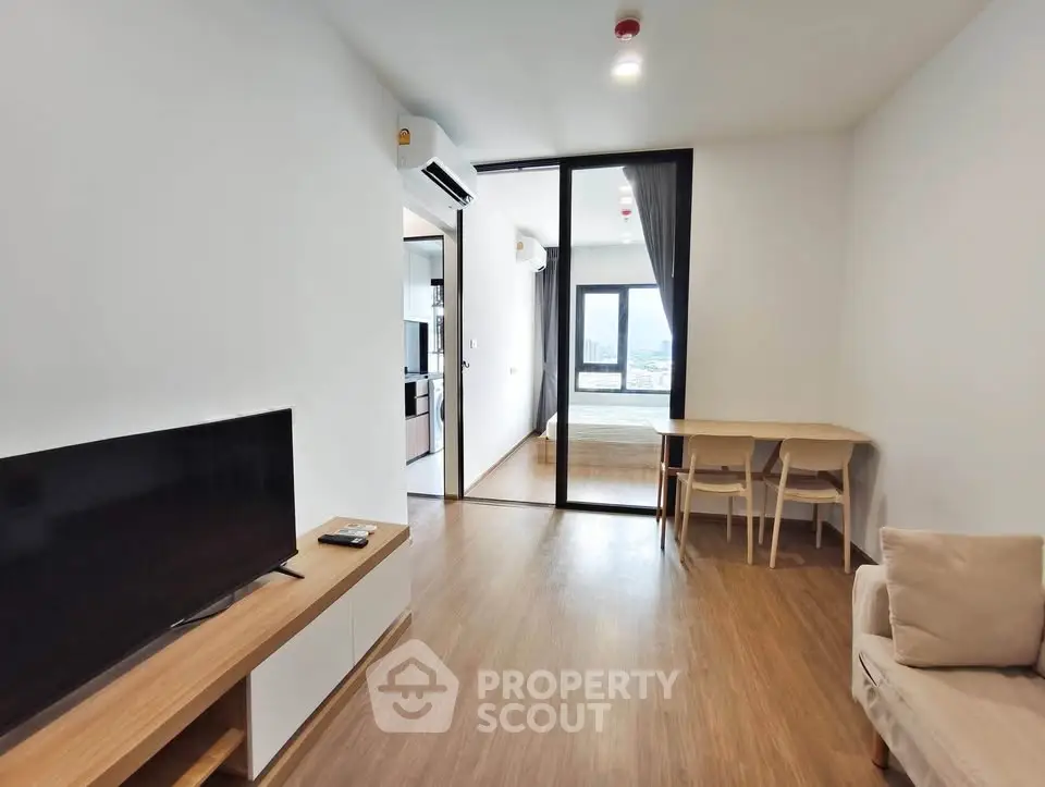 Modern living room with wooden flooring, TV unit, and view into bedroom with large window.