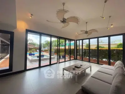 Spacious living room with large windows overlooking a pool and garden, featuring modern ceiling fans and sleek furniture.