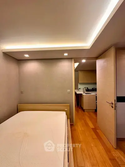 Modern bedroom with wooden flooring and recessed lighting, adjacent to a compact kitchen area.