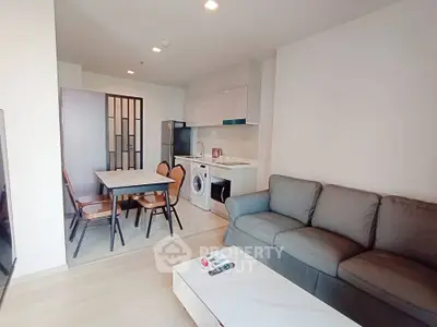 Modern living room with cozy sofa, dining area, and compact kitchen featuring washing machine and fridge.