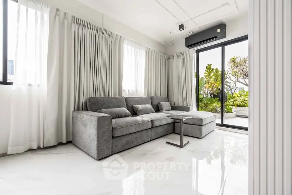 Spacious modern living room with large windows and garden view, featuring a sleek gray sofa and elegant decor.