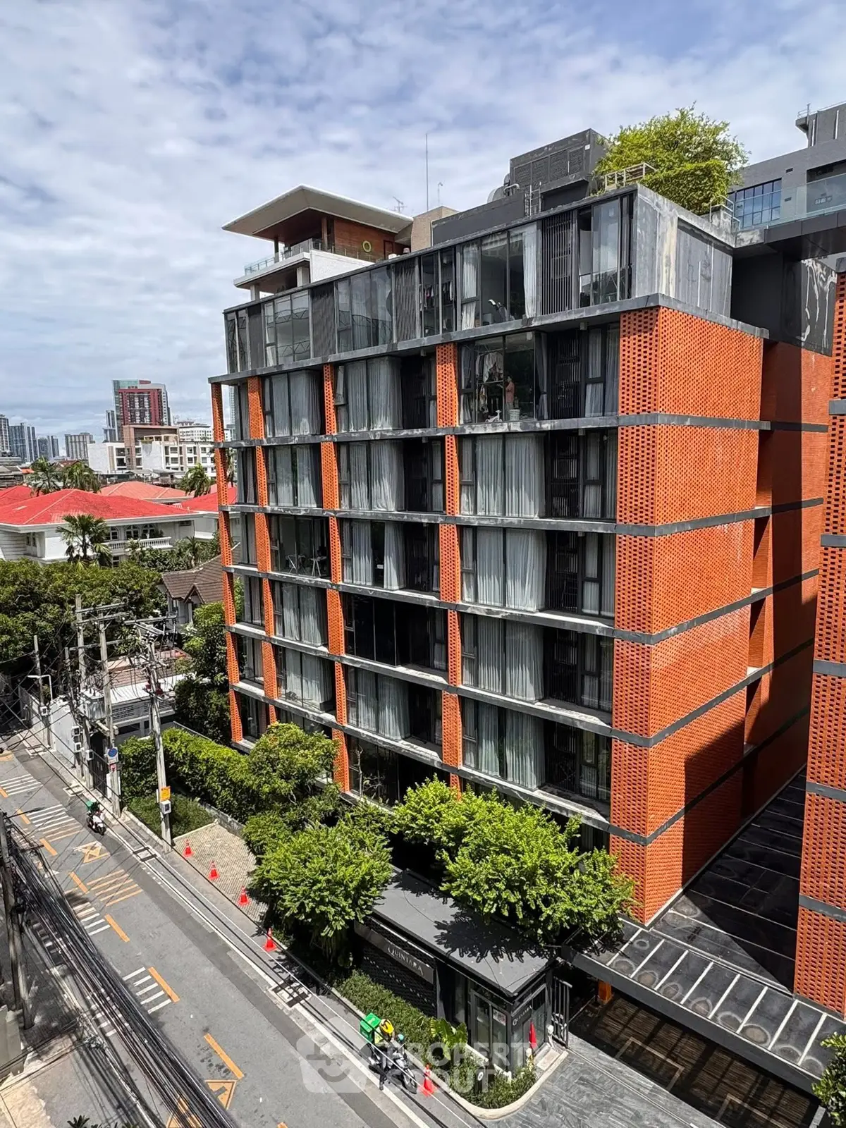 Modern urban apartment building with red brick facade and large windows, surrounded by lush greenery.